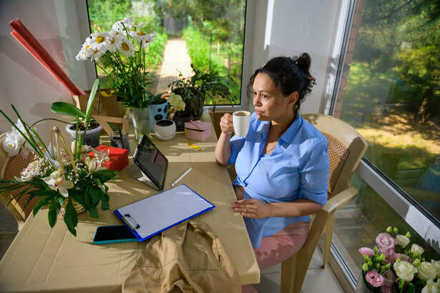 Latin American pregnant woman taking sip of coffee, sitting at table and working on clipboard in a floral design studio