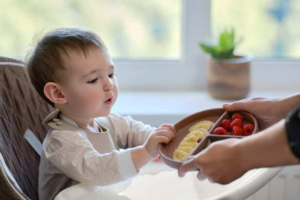 Mother gives toddler baby fruits and berries on a plate. Surprised child takes food from mom woman hands. Kid aged one year and two months