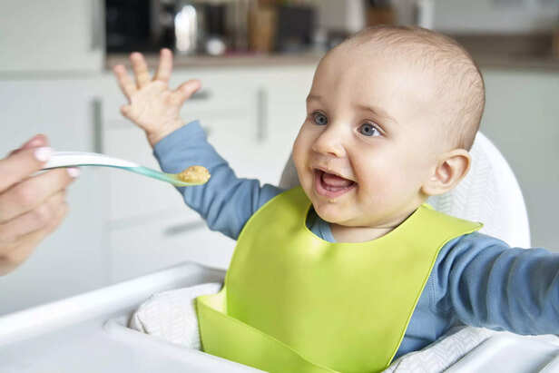 Smiling 8 month Old Baby Boy At Home In High Chair Being Fed Solid Food By Mother With Spoon