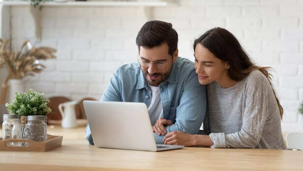 Pleasant family couple sitting at big wooden table in modern kitchen, looking at laptop for school admissions