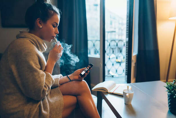 Young girl at home smoking electronic cigarette and online banking on mobile phone